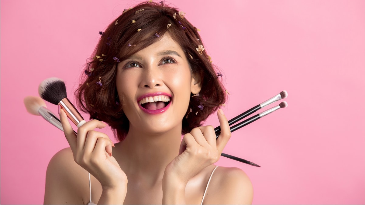 A girl is holding makeup brushes and smiling on the pink background.  