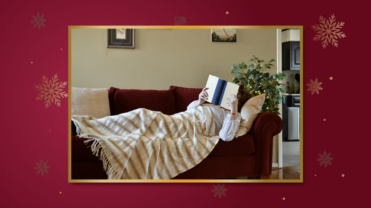 a girl lounging in her bed covered by a throw blanket is reading a book