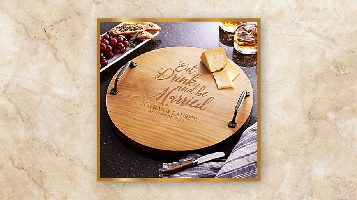 An engraved wooden chopping board with cheese on it  kept on a slab alongside berries and 2 glasses filled with a golden liquid kept as personalized wedding gifts. 
