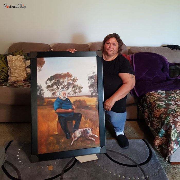 Picture of a woman sitting on a couch holding a portrait that shows a man with a dog sitting in on a wood