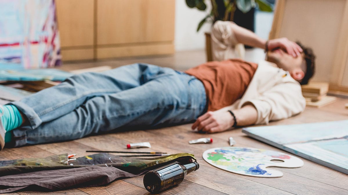 A man tired lying down next to his paint palette and canvas, with a hand over his head. He seems to be tired and in distress. 