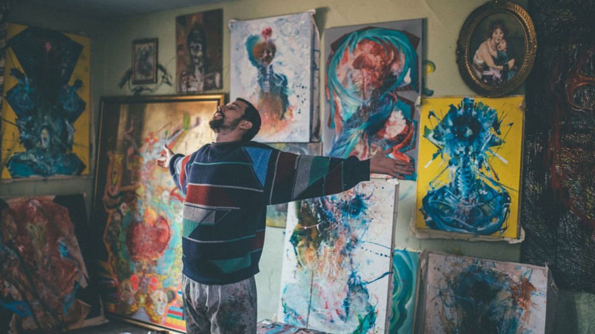 a man standing surrounded with paintings and traditional art, with closed eyes and open arms.