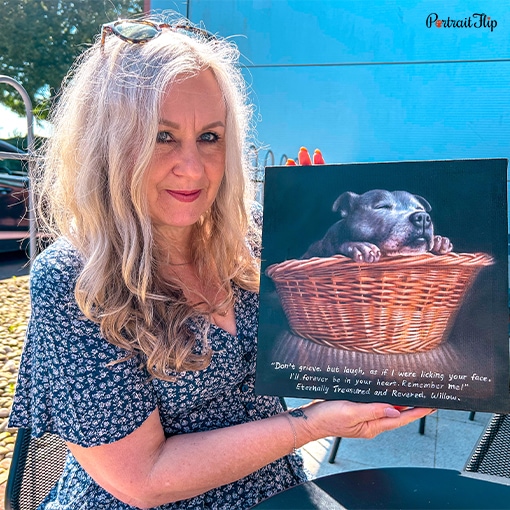 Picture of an old woman holding a pet portrait that shows her dog in a wooden basket