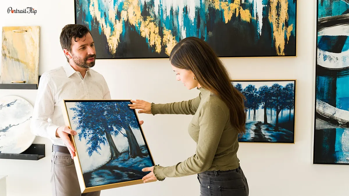 A man holding a painting of trees colored in blue which a woman is looking at